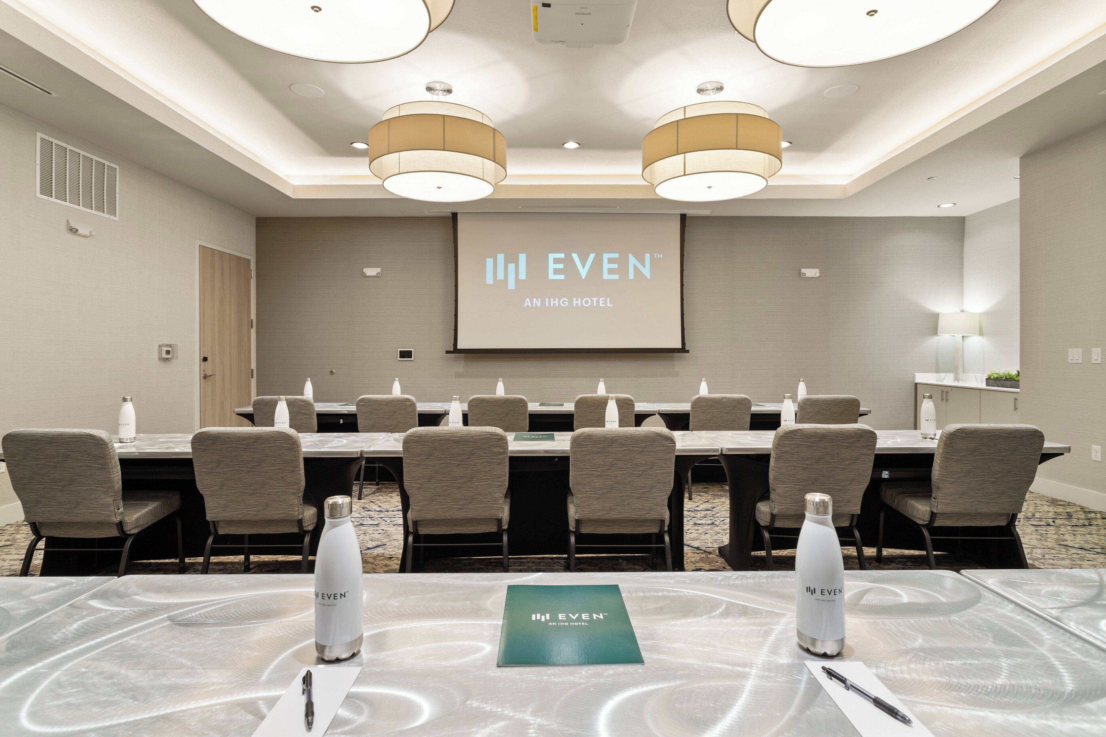 A conference room with a long table, chairs, water bottles, and a presentation screen displaying "EVEN AN IHG HOTEL".