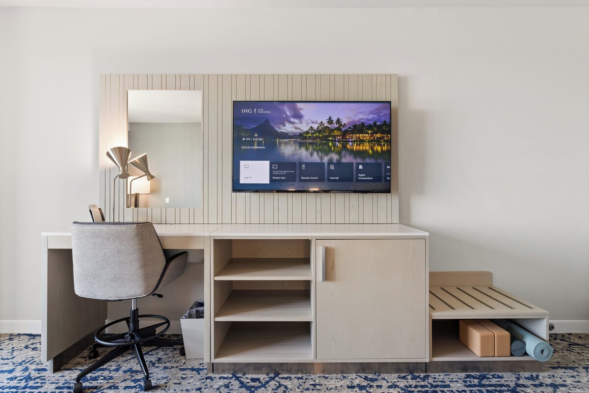 Une chambre d’hôtel moderne avec un bureau en bois, une chaise, un téléviseur à écran plat fixé au mur et un petit lit doté d’une tête de lit en bois.