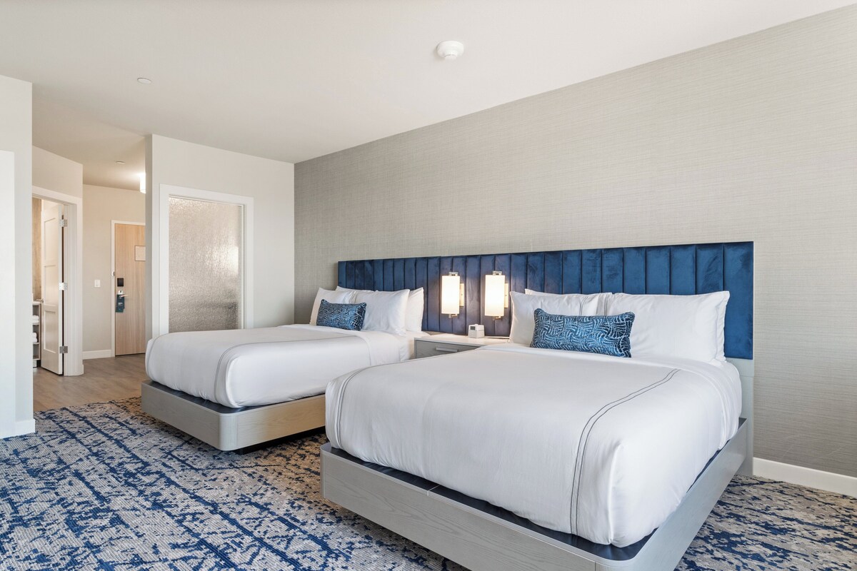 Une chambre d’hôtel avec deux lits, une tête de lit bleue, du linge de lit blanc et un tapis à motifs.