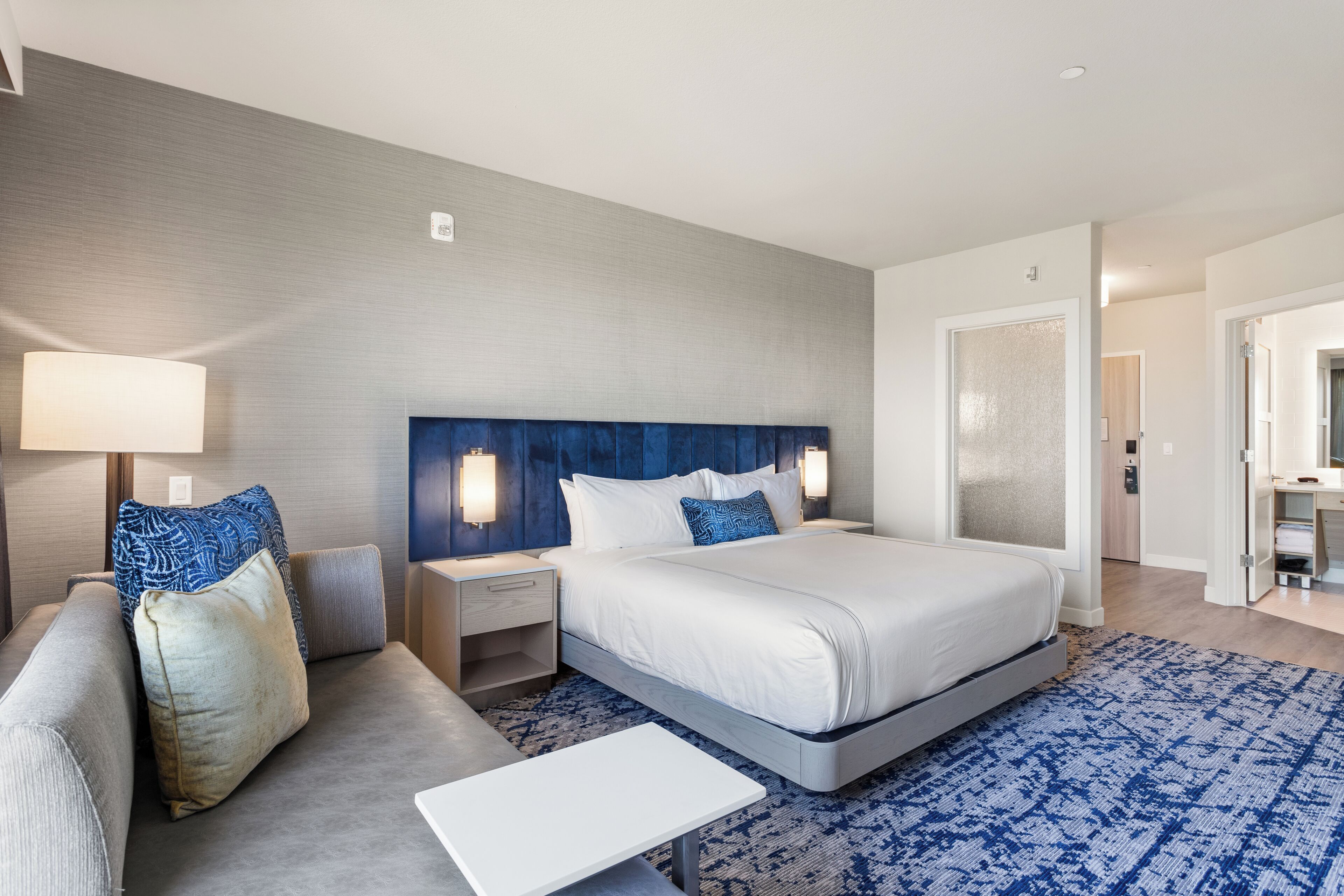 Una habitación de hotel con una cama grande, un sofá, una mesita de noche y una lámpara.
