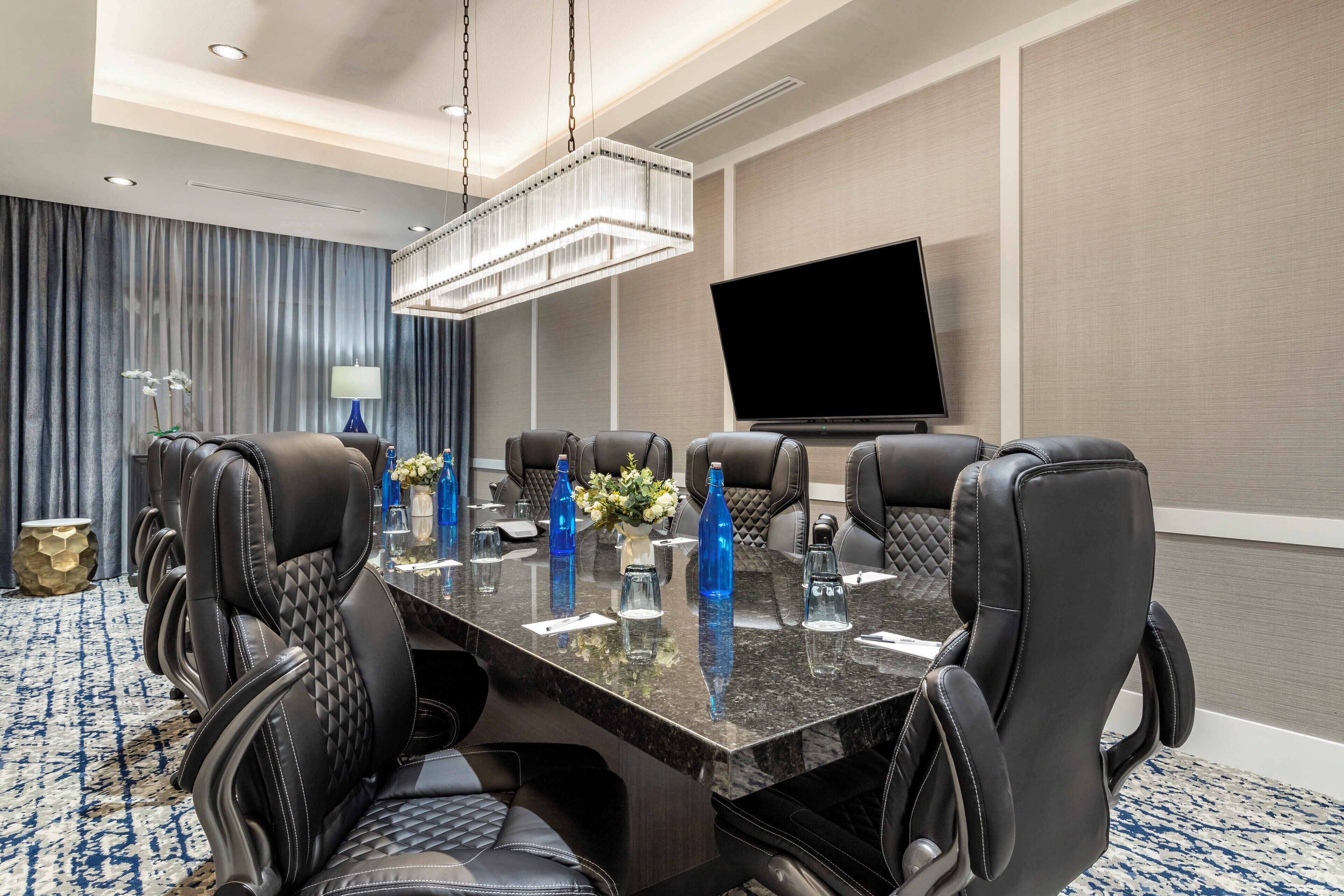 A conference room with a large table, black leather chairs, a flat-screen TV, and a chandelier.