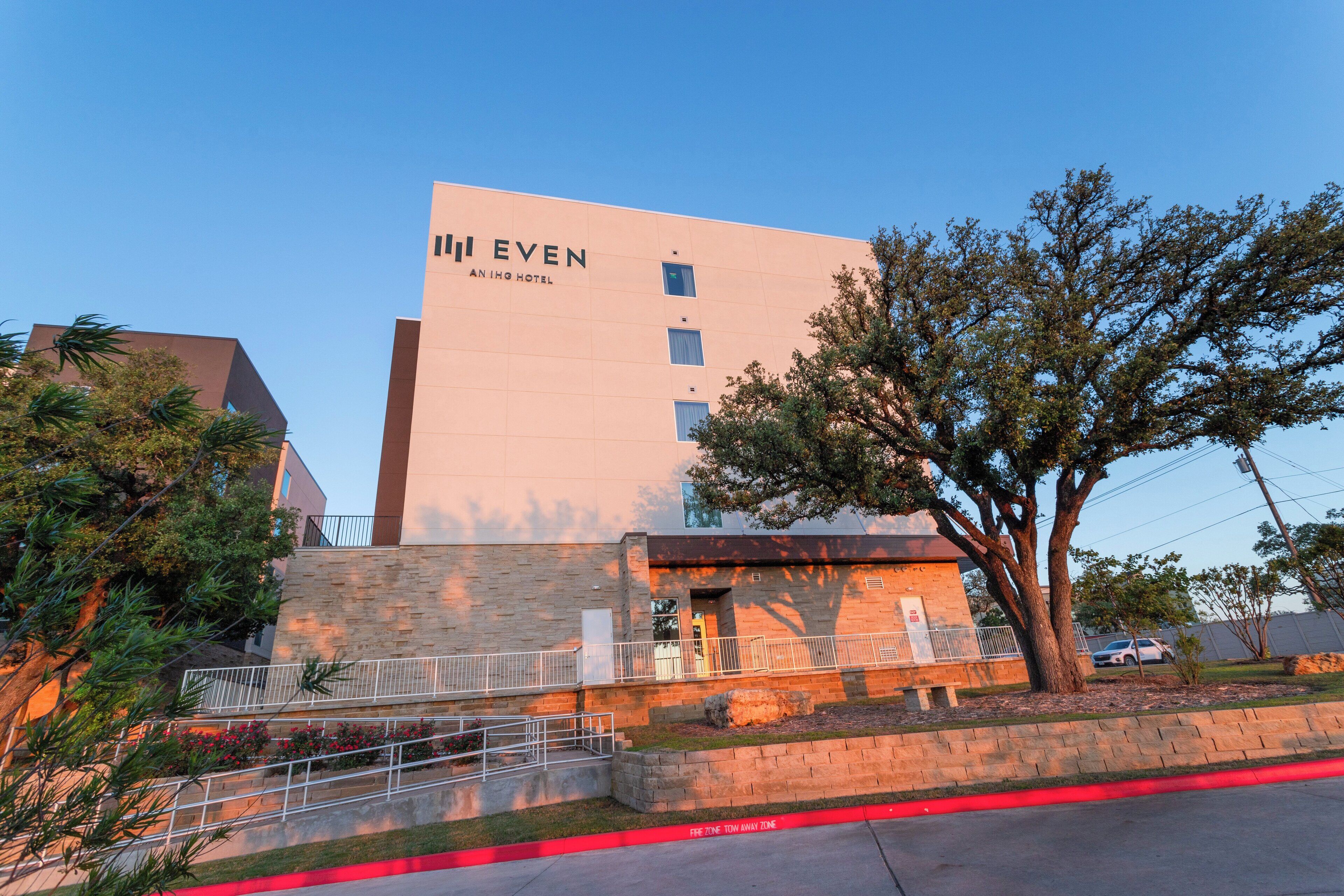 A hotel named 'II EVEN' with a modern design, featuring a prominent entrance, a landscaped area with trees, and a clear sky.