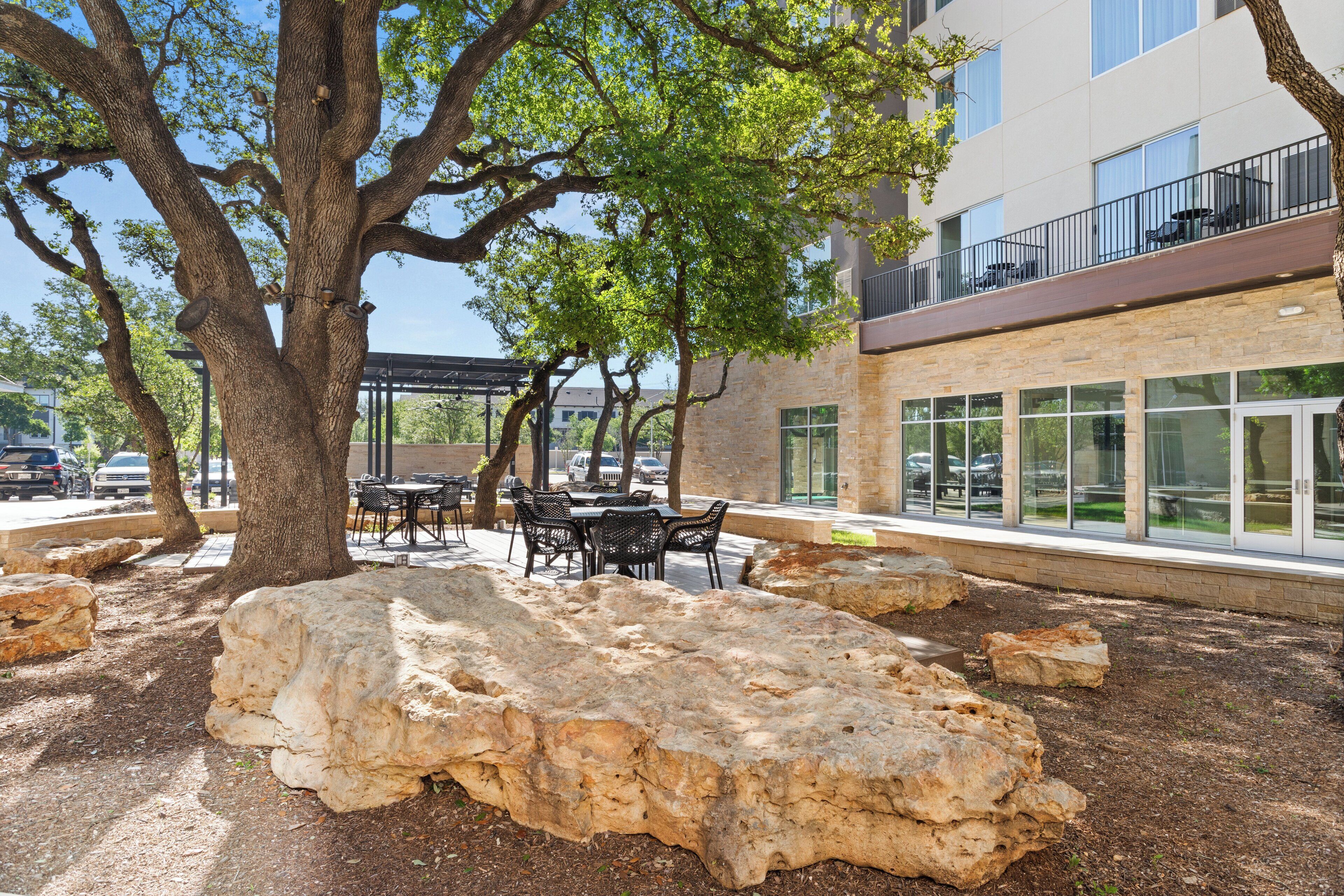An outdoor seating area with tables and chairs, a large rock formation, and a building with large windows and balconies.