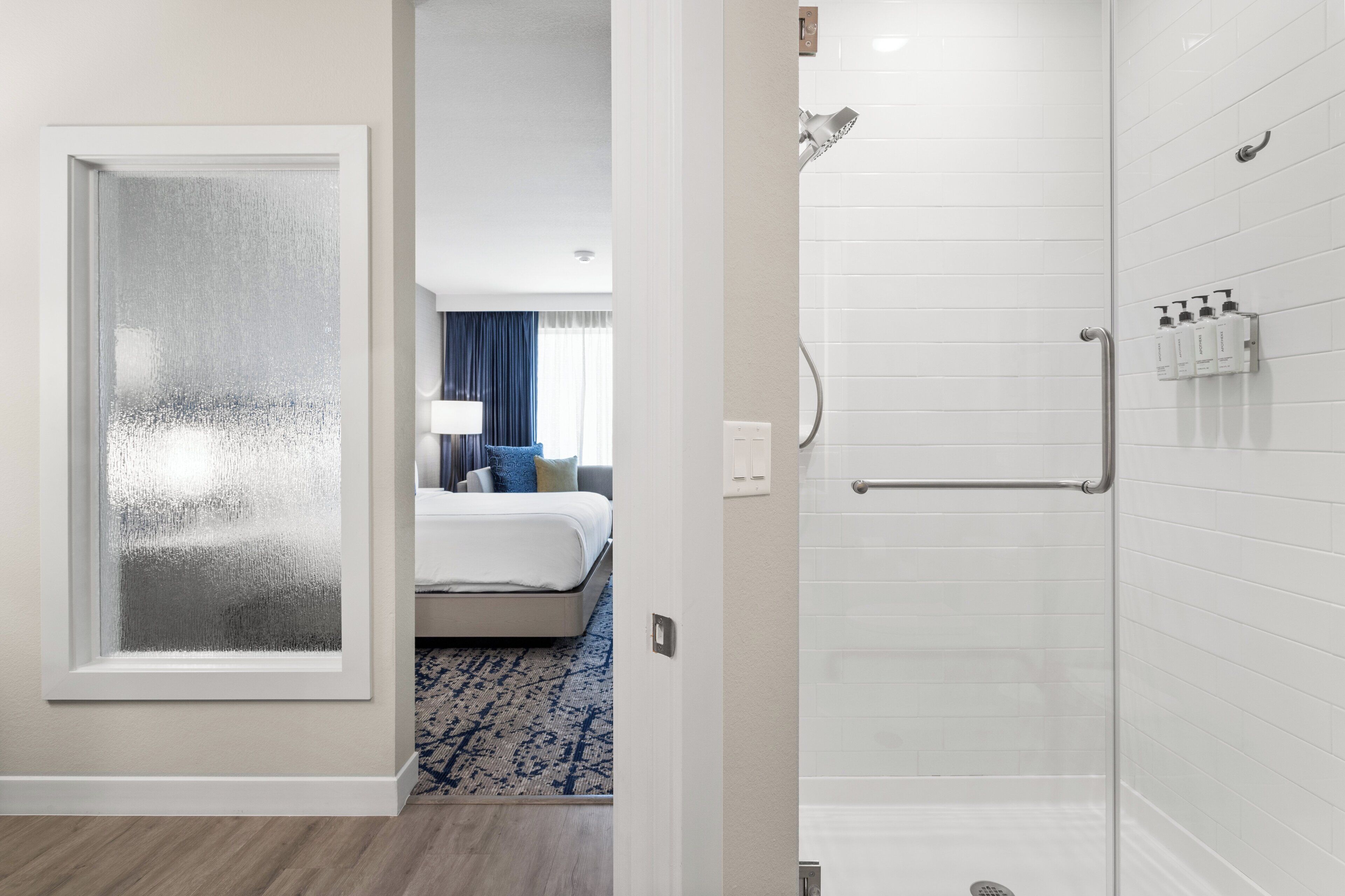 A hotel room with a glass door leading to a bedroom, a shower area with white tiles, and a visible bathroom counter with toiletries.
