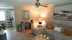 A living room with a grey sofa, a coffee table, a ceiling fan, a lamp, a painting, and a fireplace. - The Cozy Corner, Family Friendly, Game Room + WiFi (Woodward)