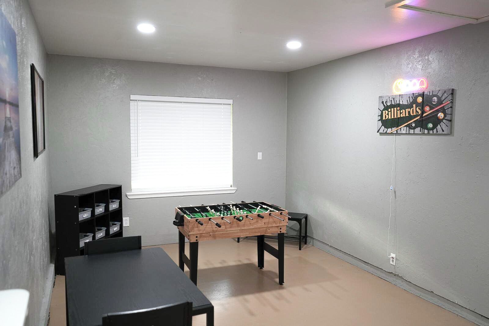 A room with a foosball table, a billiards table, and a window with blinds.