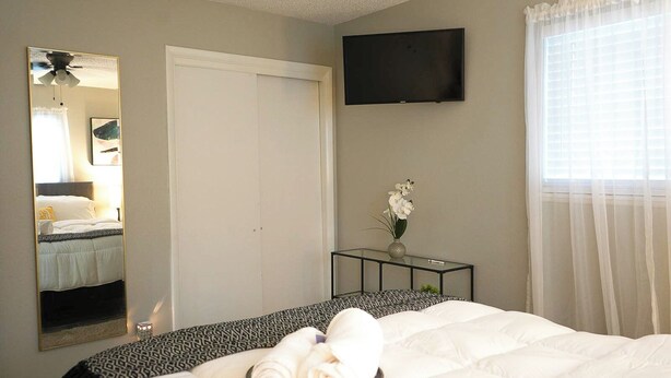 A hotel room with a bed, a TV mounted on the wall, a bedside table with a vase of flowers, and a window with sheer curtains. - The Cozy Corner, Family Friendly, Game Room + WiFi (Woodward)
