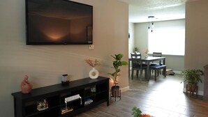 A modern living room with a flat-screen TV mounted on the wall, a black entertainment unit with decorative items, a dining area with a table and chairs, and a potted plant. - The Cozy Corner, Family Friendly, Game Room + WiFi (Woodward)
