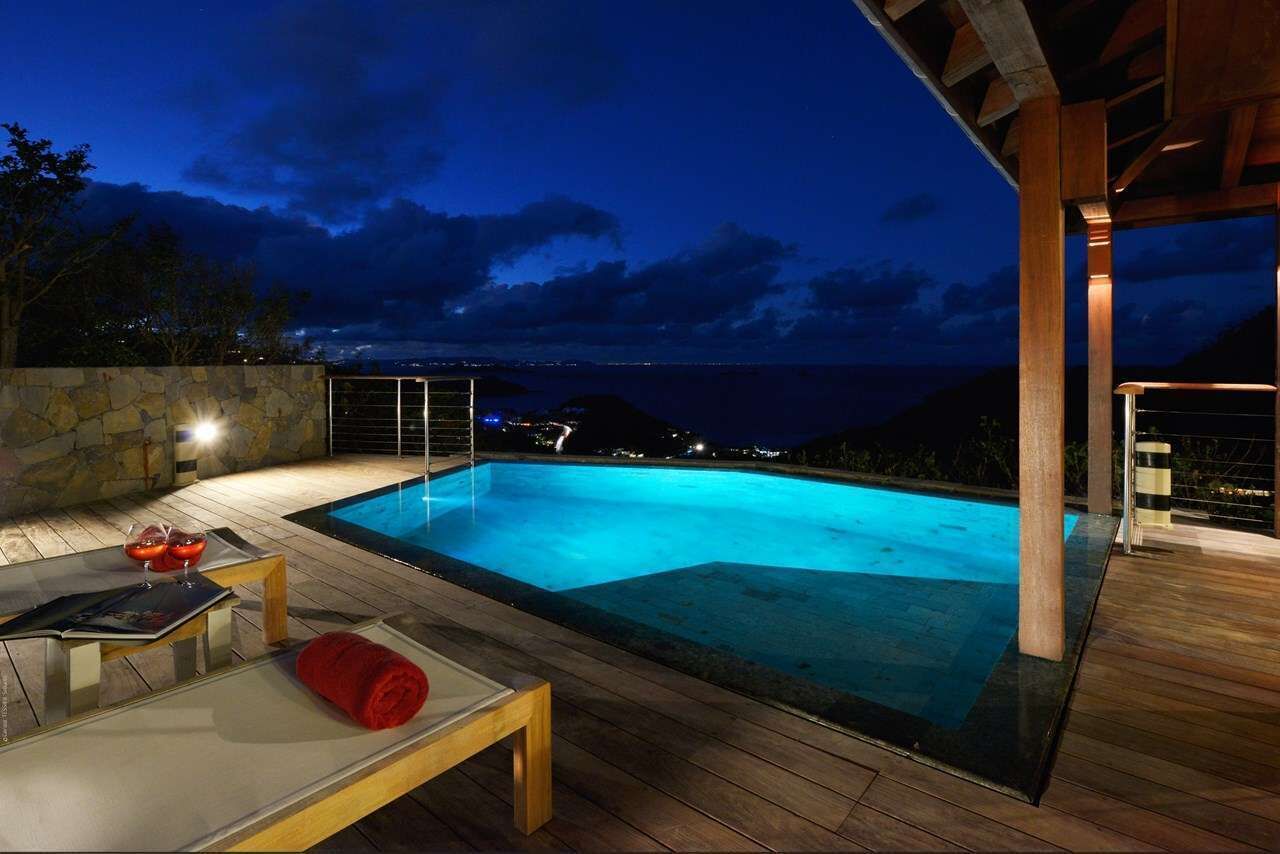 A swimming pool with a stone wall and wooden deck, a lounge area with a red towel, and a view of the ocean at night.