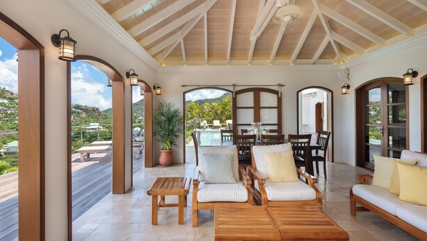 Un espacio de estar al aire libre techado con mobiliario de madera y vistas a una piscina y montañas.