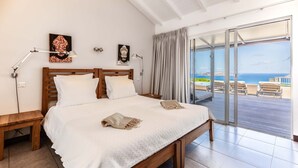 A bedroom with a wooden bed, white linens, a nightstand, a lamp, and a view of the sea through a sliding glass door.