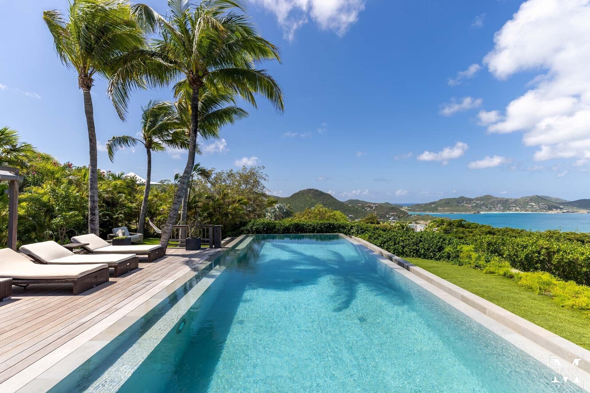 A swimming pool with lounge chairs, palm trees, and a view of the ocean and hills.