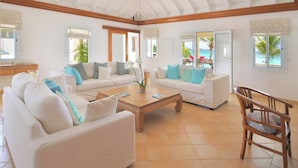 Amplia sala de estar con dos sofás blancos, una mesa de centro de madera y una silla de madera. La habitación tiene ventanas amplias con vistas a palmeras y al océano.