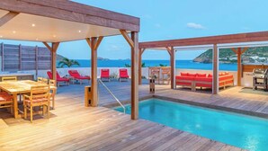 Una terraza de madera con piscina, sillas reclinables rojas y una mesa.