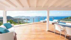 Eine Holzterrasse mit einem weißen Sofa und Stühlen, einem Tisch und Blick auf das Meer und die Hügel.