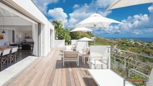 Un patio exterior moderno con suelo de madera, mobiliario blanco y grandes sombrillas que dan sombra.