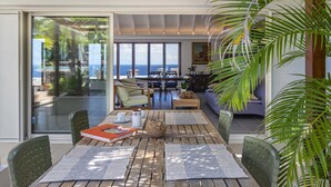 Amplia sala de estar con un gran ventanal corredera que da a un balcón con vistas al océano. En el interior, se encuentra una mesa de comedor de madera con sillas, un sofá y una mesa baja.