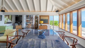 Amplia sala de estar con techo alto, un ventanal con vista al mar, una mesa de comedor con sillas y una estantería.