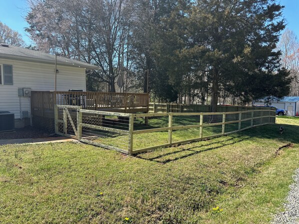 A fenced outdoor area with a wooden deck, a house in the background, and trees.
