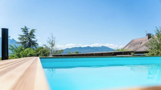 A clear blue swimming pool with a wooden deck in the foreground, a building with a sloped roof in the background, and mountains in the distance.