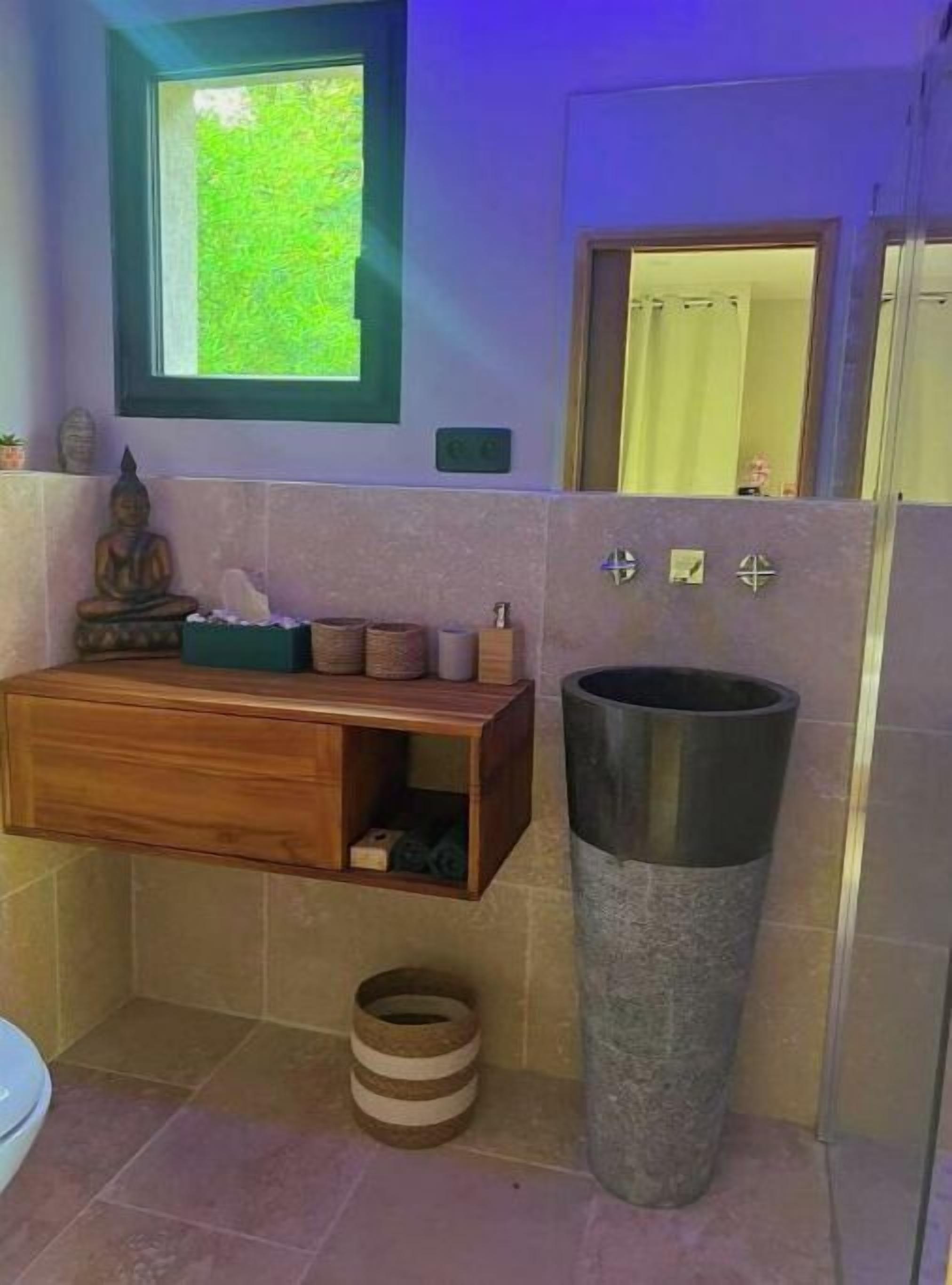 A bathroom with a wooden vanity, a large cylindrical sink, and a window with a view of greenery.
