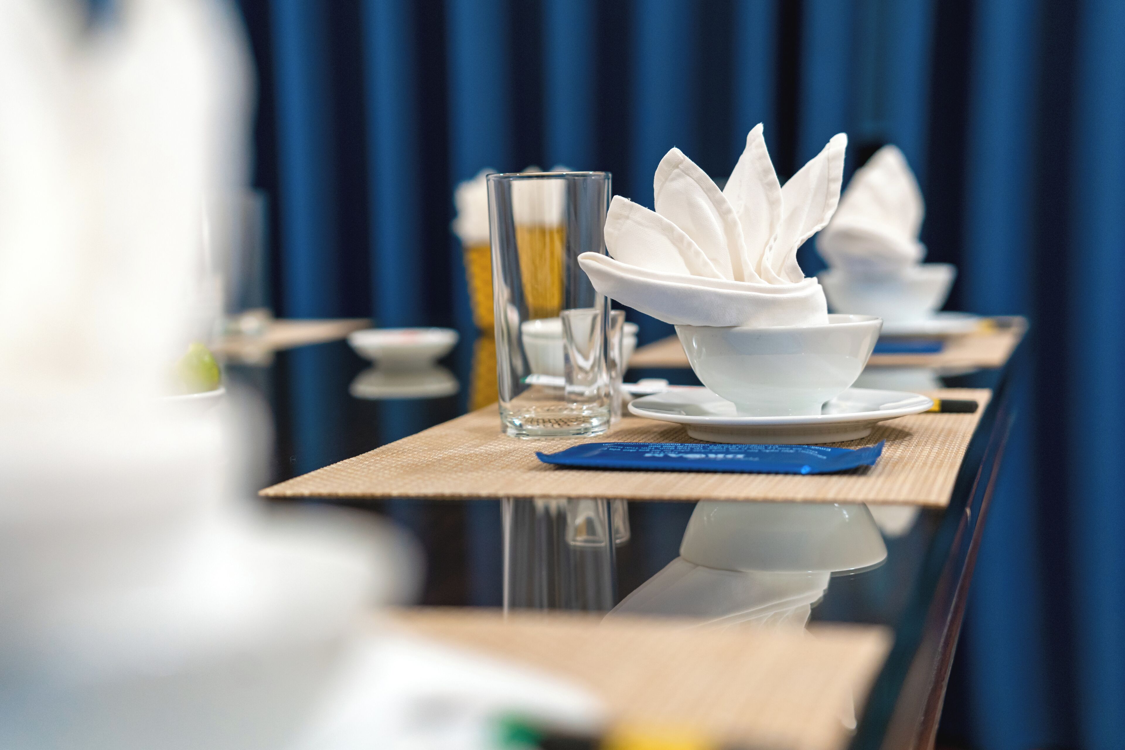 A dining table with white dishes and a glass of water, set against a blue curtain background.