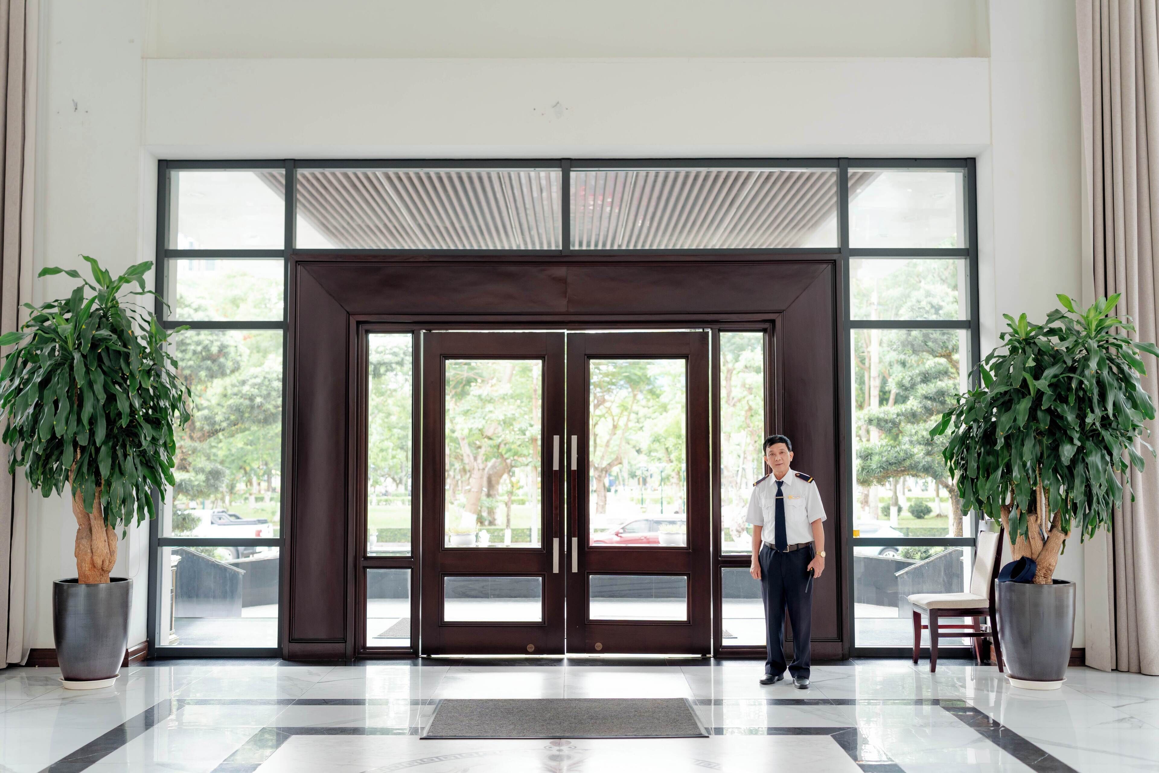 A man standing in front of a double door with a view of trees and a parking lot. There are two potted plants on either side of the door.