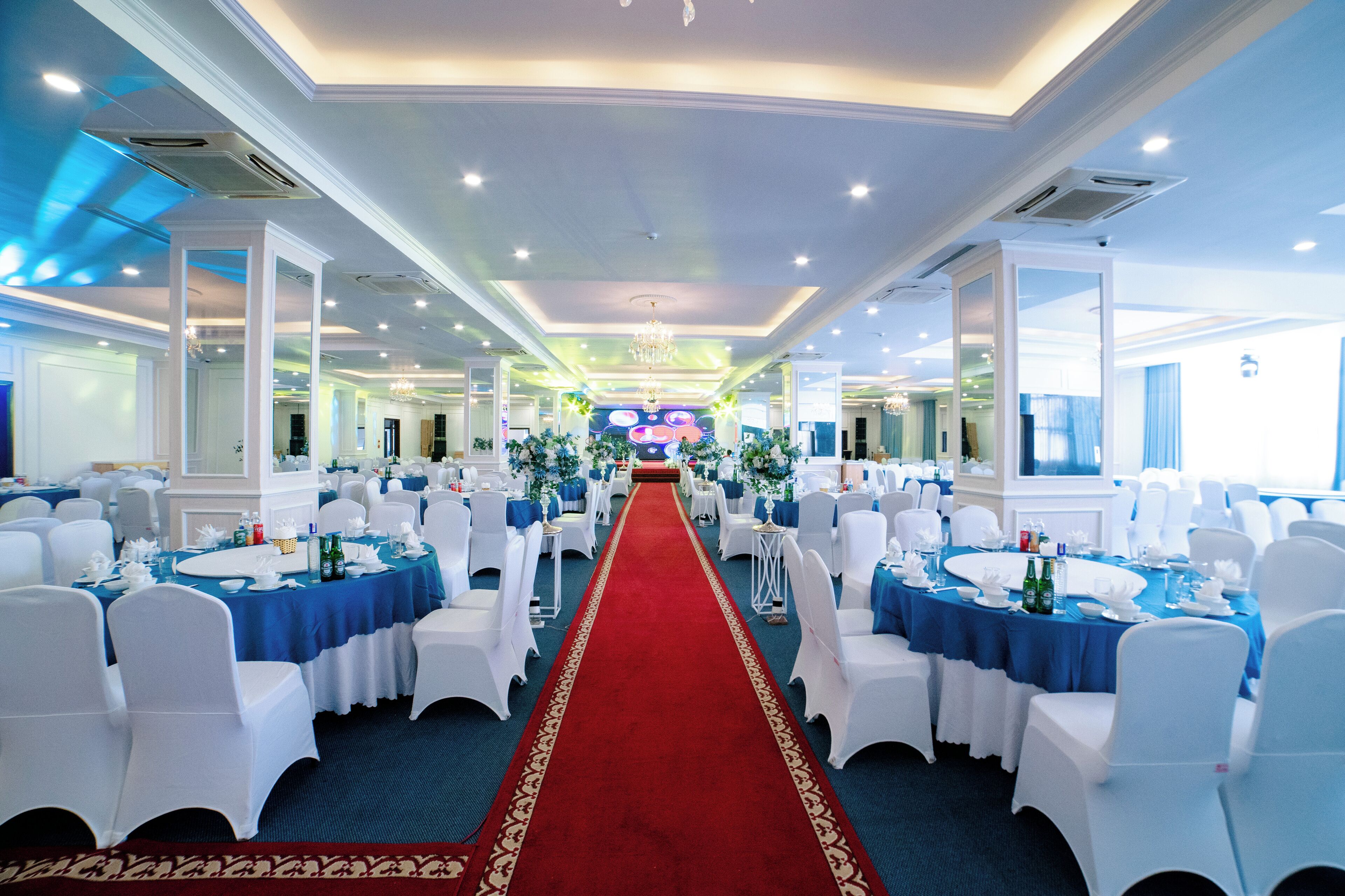 A banquet hall with white chairs, blue tablecloths, and a red carpet leading to the front.