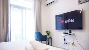 A hotel room with a bed, a TV mounted on the wall displaying the YouTube logo, a blue chair, and a small table with a plant. - Beautiful Suite @Embassy Gardens (Accra)