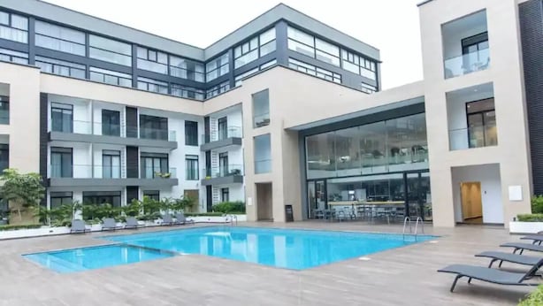 A modern building with a swimming pool, lounge chairs, and a glass facade. - Beautiful Suite @Embassy Gardens (Accra)