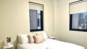 Une chambre d’hôtel avec un lit, deux fenêtres, une table de chevet avec une plante et une serviette roulée posée sur le lit.