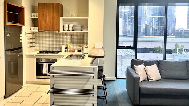 Une cuisine moderne dotée d’un four et d’un micro-ondes encastrés, un coin repas avec une table en bois, et un canapé gris offrant une vue sur la ville grâce à de grandes fenêtres.
