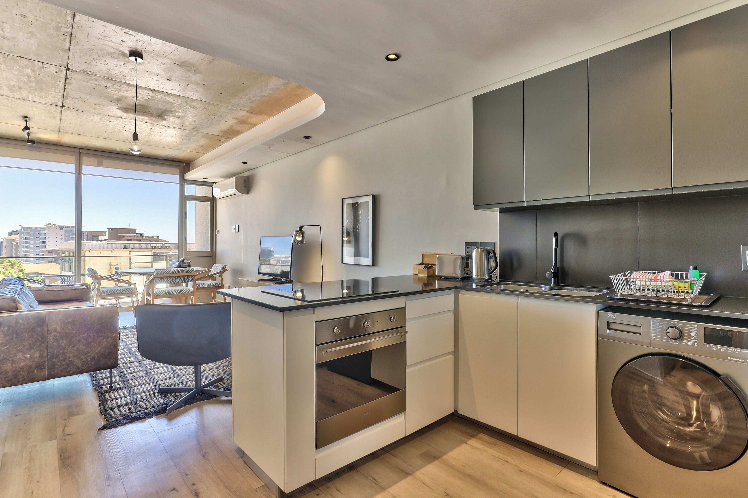 A modern kitchen with a built-in oven, dishwasher, and washing machine. There is a dining area with a table and chairs, and a large window offering a view of the city.