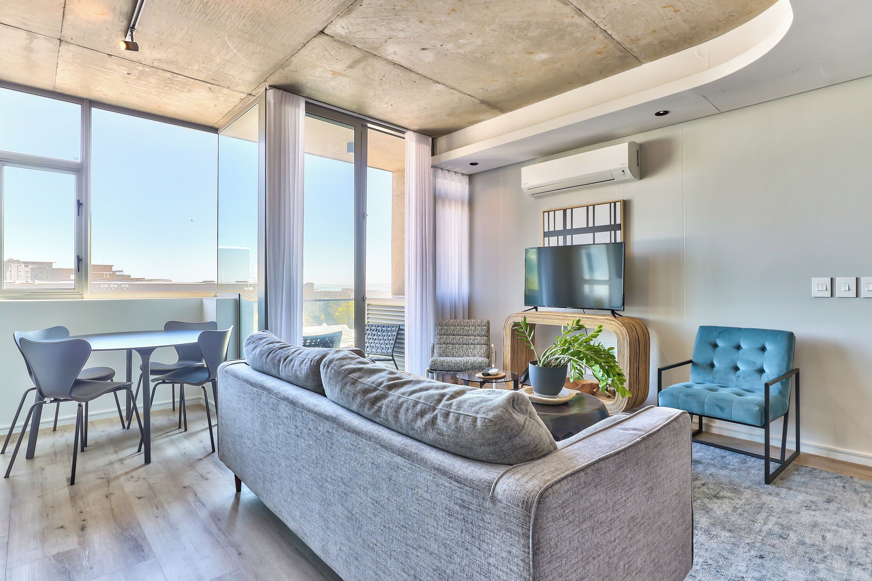 Un salon moderne avec un canapé gris, un fauteuil bleu, une table à manger avec des chaises et une grande fenêtre avec des rideaux.