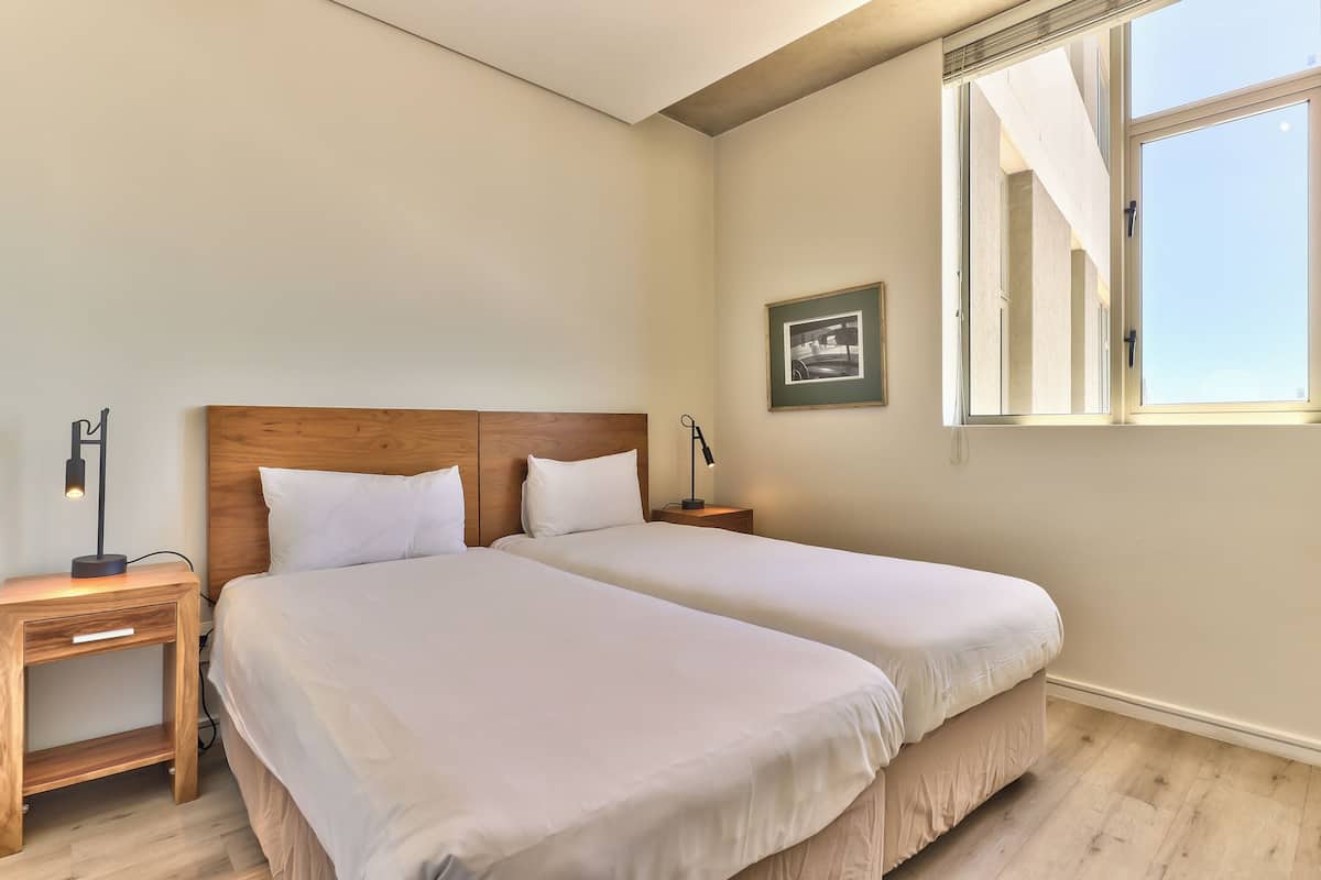 Une chambre à coucher avec deux lits, des têtes de lit en bois, des tables de chevet avec des lampes, un tableau encadré au mur et une fenêtre avec des rideaux.