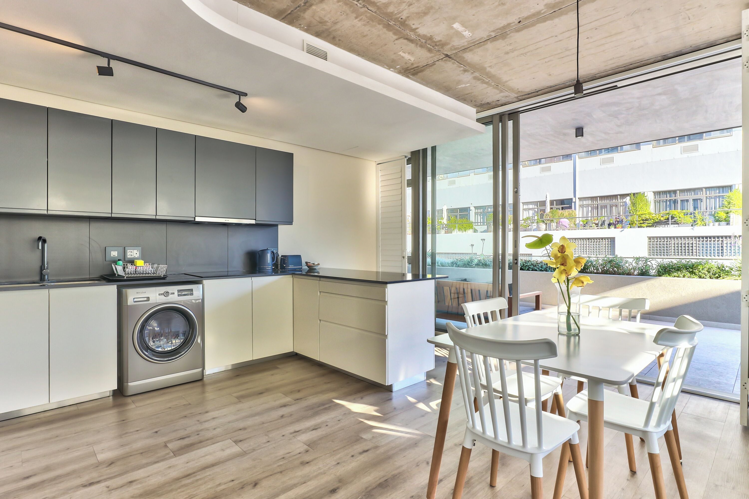 A modern kitchen with a built-in washer, large windows, and a dining area with white chairs and a table.