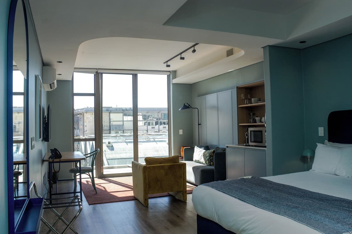 Une chambre d’hôtel moderne, dotée d’un grand lit, d’un bureau avec une chaise, d’un petit canapé et offrant une vue sur la ville grâce à une grande fenêtre.