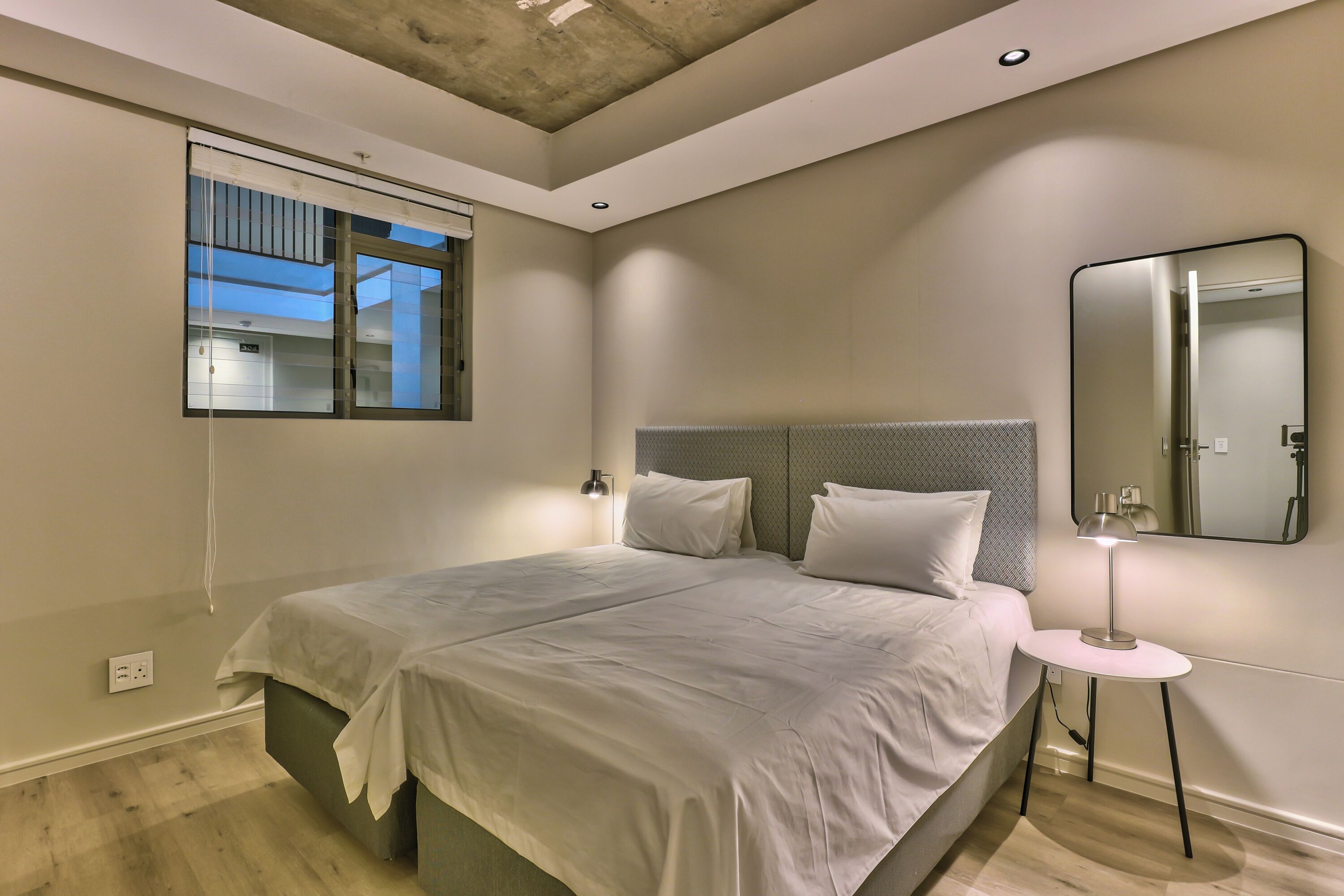 A modern bedroom with a large bed, a bedside table with a lamp, a window with blinds, and a mirror on the wall.