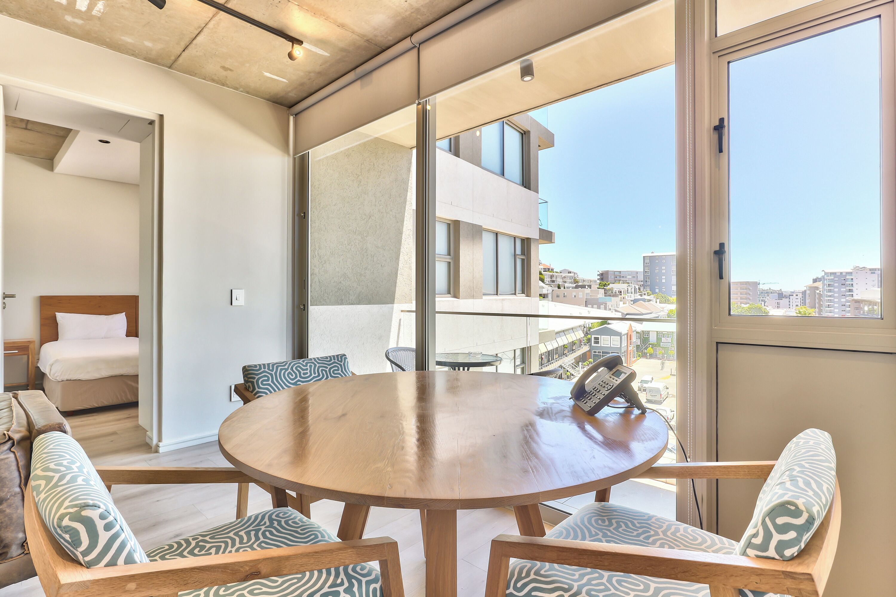 Quarto moderno com uma mesa redonda de madeira, sofás estampados, uma cama e grandes janelas com vista para a cidade.