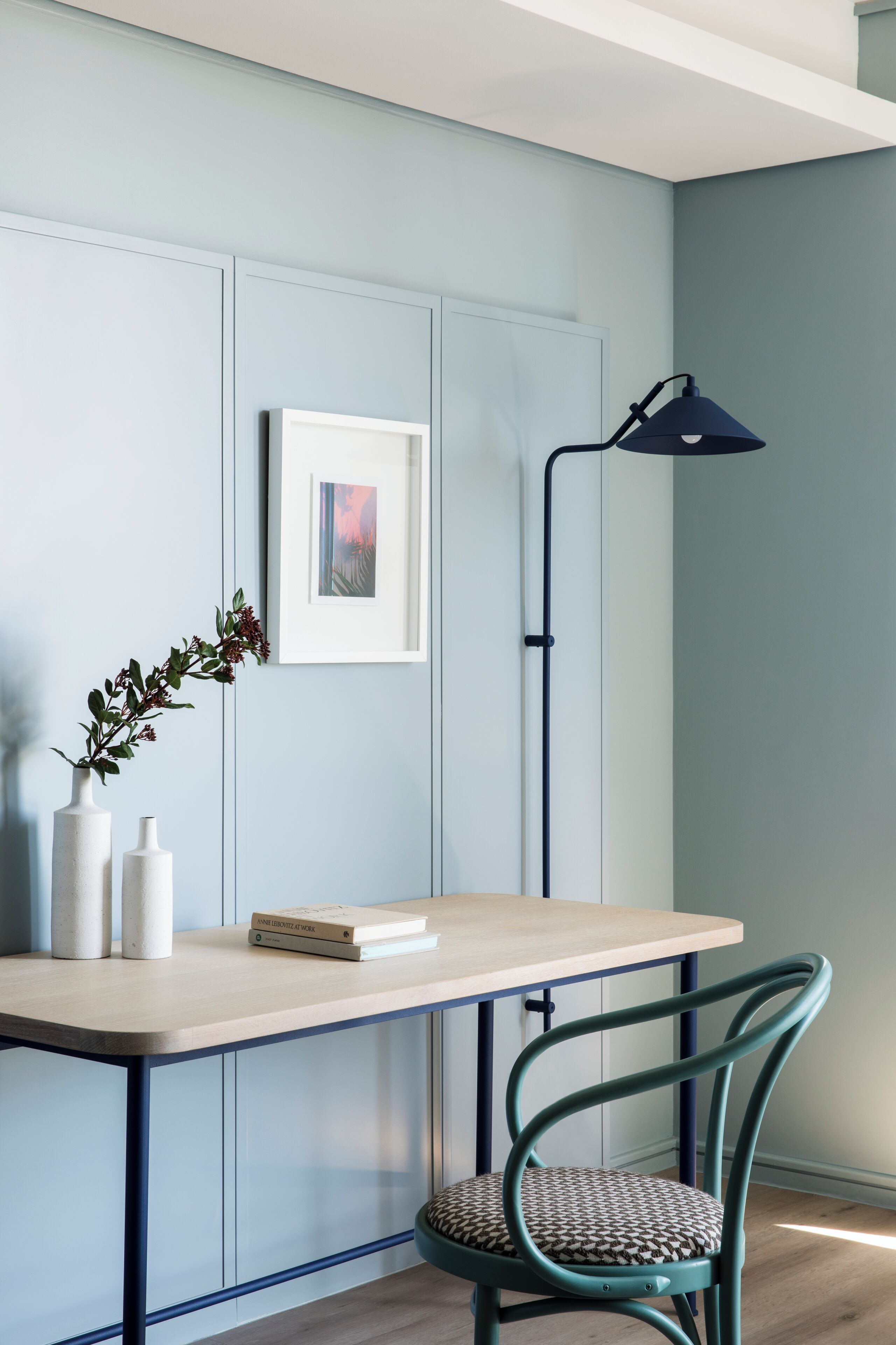 A room with a light blue wall, a framed picture, a table with two vases and a book, a teal chair, and a floor lamp.