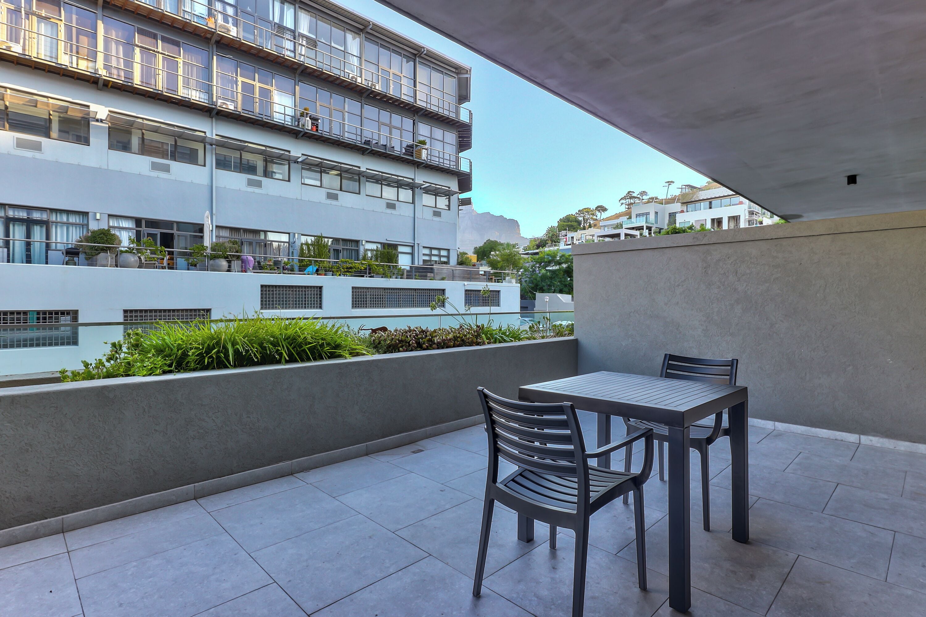 Varanda externa moderna com mesa e cadeiras, com vista para uma área residencial com um edifício de vários andares e vegetação.