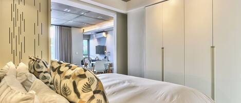 A modern hotel room with a large bed, a wardrobe, and a view into the dining area.