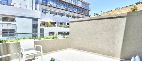 Een terras buiten met witte meubels, een kleine tafel met een plant en uitzicht op een modern gebouw met balkons.