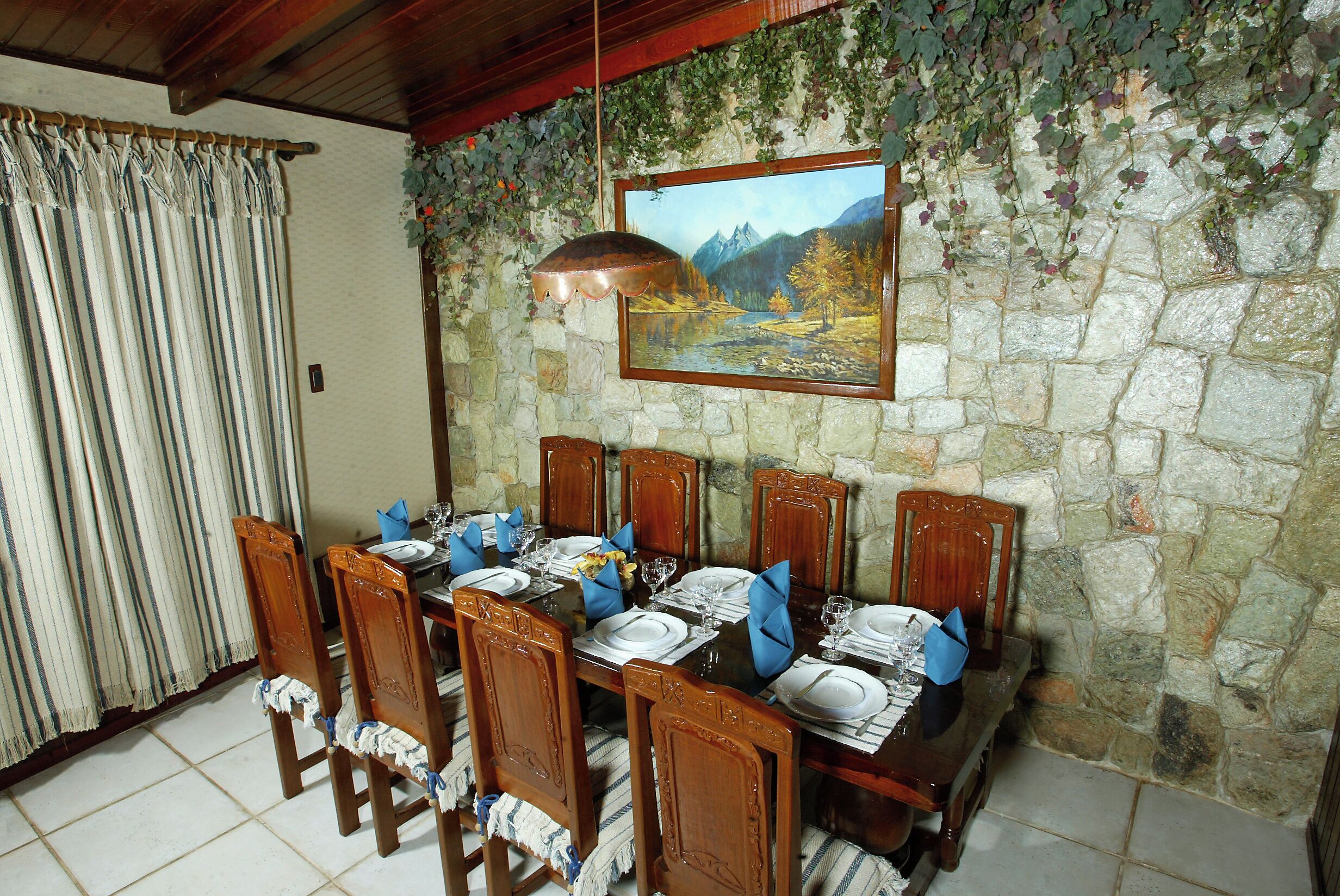 A dining area with a stone wall, wooden chairs, a table set for six, and a painting above the table.