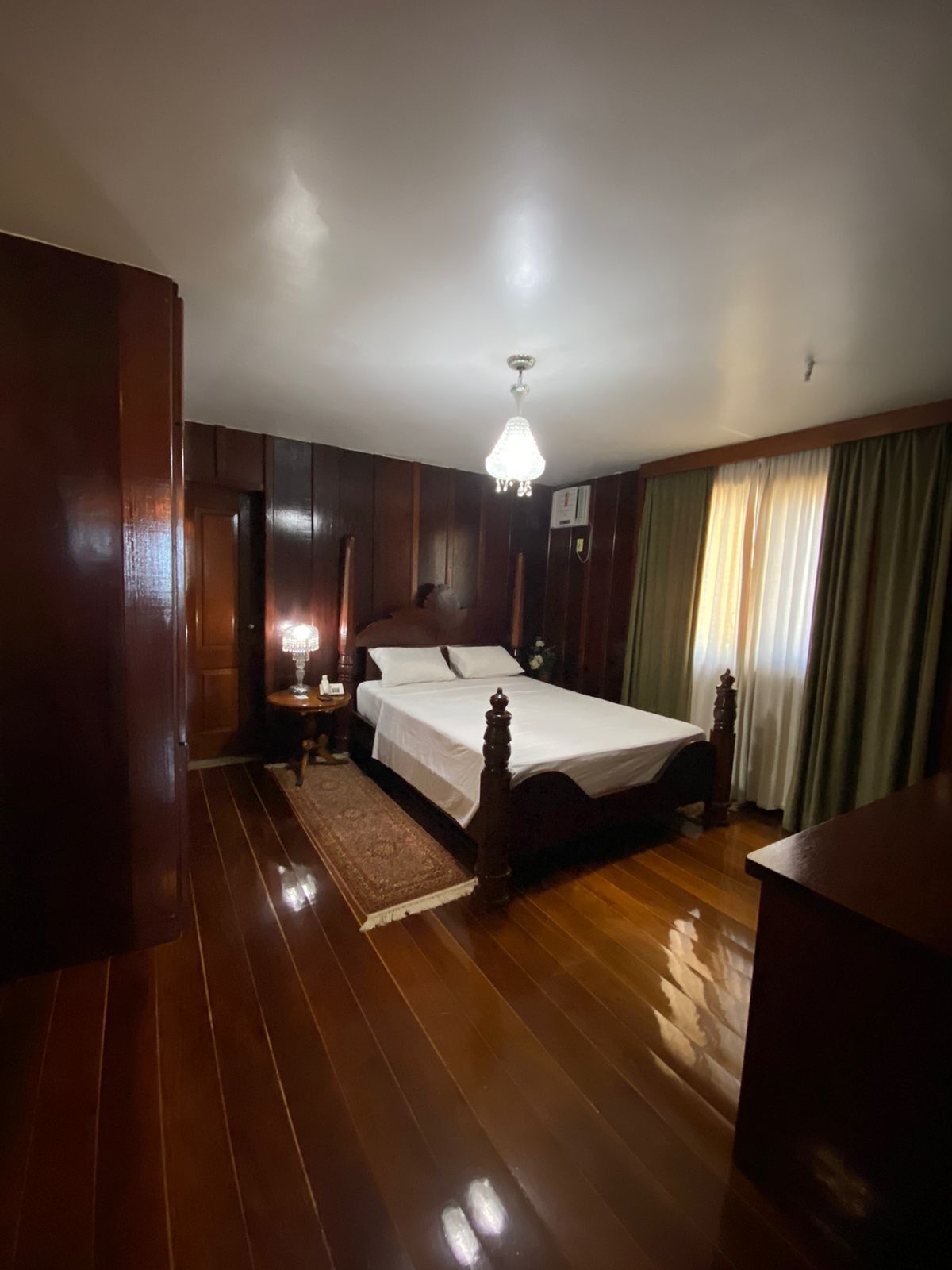 A bedroom with a wooden floor, a bed with a white bedspread, a nightstand, a lamp, and a chandelier.