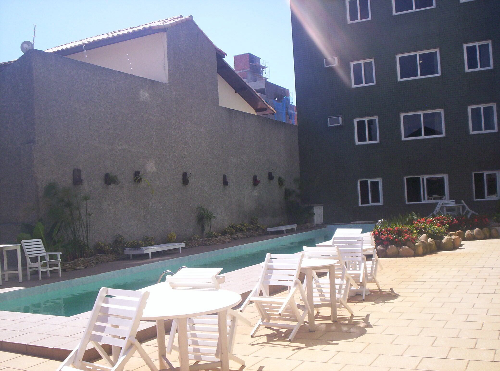 Un área de piscina con sillones y mesas, un pequeño jardín con plantas y flores, y un edificio con múltiples ventanas y techo de tejas.