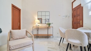 A room with a wooden sofa, a dining table with chairs, a small side table with a lamp, and a wall-mounted mirror.