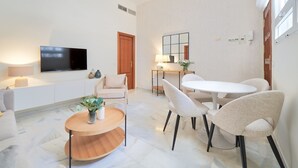 A modern living room with a flat-screen TV, a sofa, a round coffee table, and a dining area with a white table and chairs.