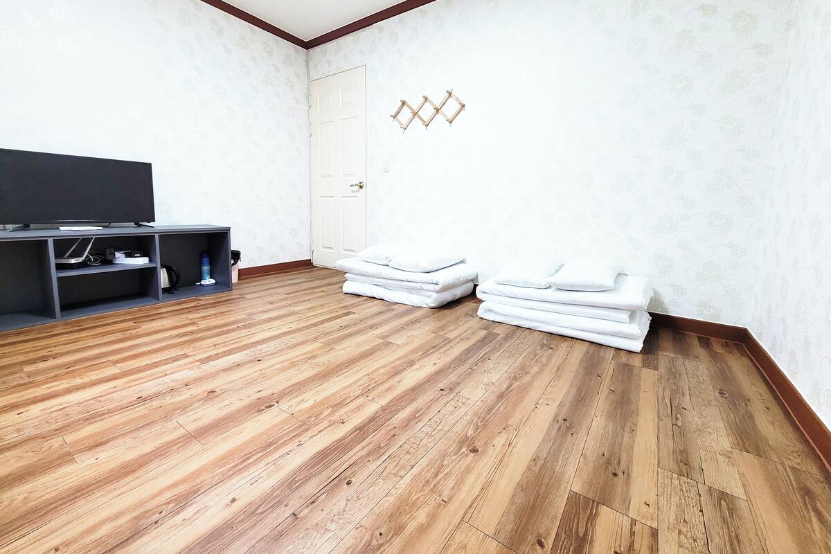 A room with wooden flooring, a flat-screen TV on a stand, and neatly stacked white pillows.