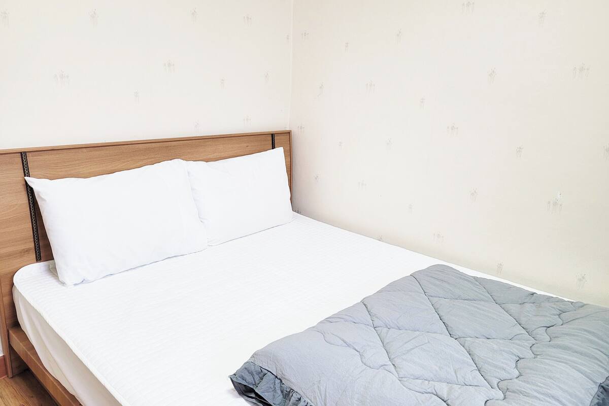 A neatly made bed with white bedding and a grey blanket, a wooden headboard, and a plain wall with a subtle pattern.